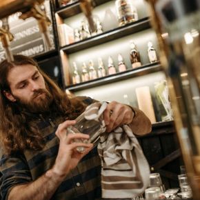 Bearded bartender cleaning a glass at a bar with various bottles displayed on shelves in the background near LOCAL Boise apartments.