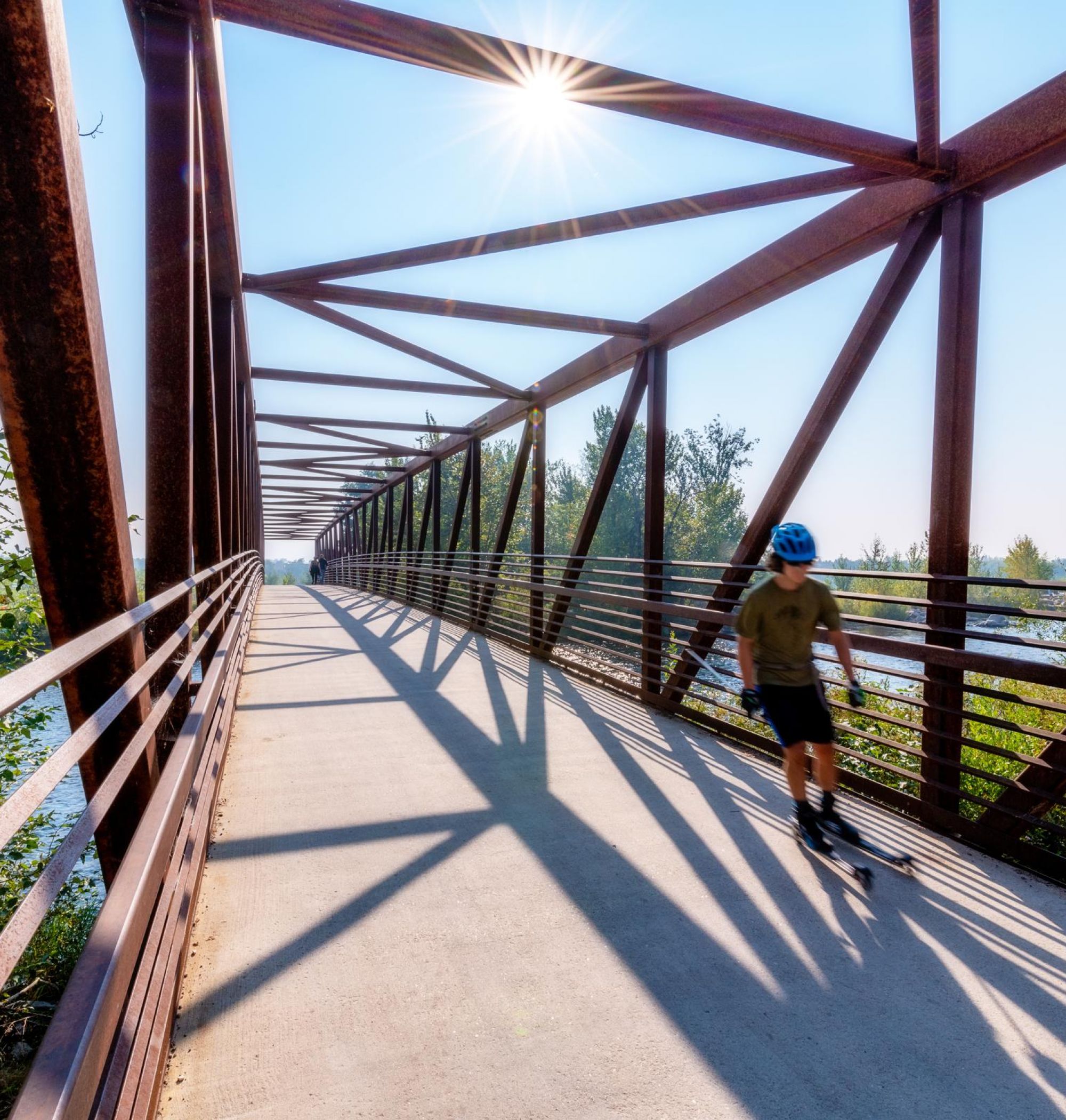 A person rollerblading on a sunlit, metal-framed bridge with greenery and river visible through the railings near LOCAL Boise apartments.