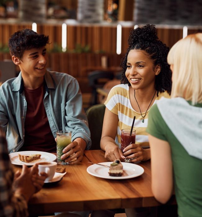Four friends smiling and chatting at a cafe table, enjoying drinks and cake near LOCAL Boise apartments.