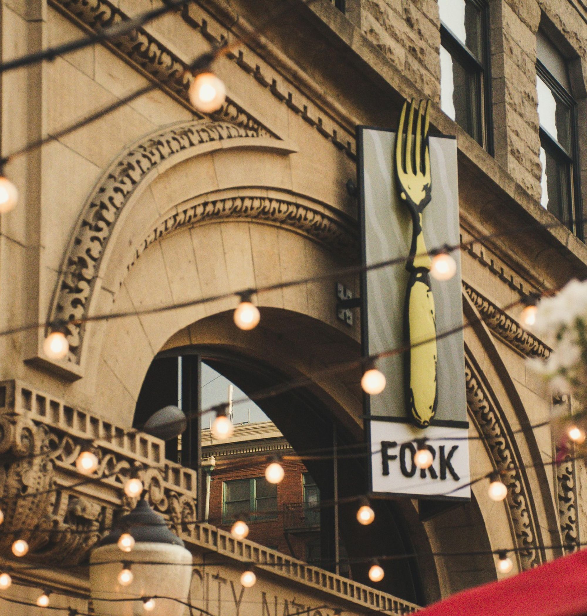 Building facade with ornate arch, lit string lights, sign with a large fork, and hanging flower basket near LOCAL Boise apartments.
